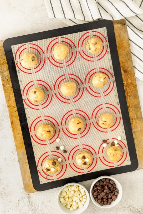 These soft and chewy Chocolate Chip Walnut Cookies have the perfect balance between sweet and nutty with two kinds of chocolate chips. A baking sheet lined with a silicone mat holds 12 unbaked cookie dough balls, some with chocolate chips pressed into the top. Nearby are three small bowls filled with white chocolate chips and regular chocolate chips.