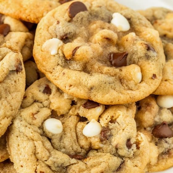 A close-up of a plate piled with freshly baked chocolate chip walnut cookies, featuring both white and dark chocolate chips, on a white background.