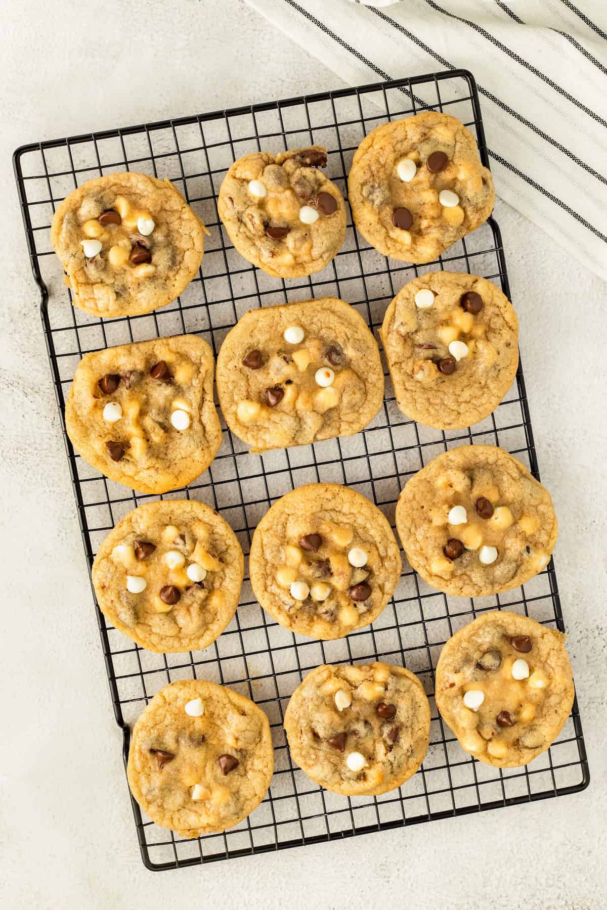 These soft and chewy Chocolate Chip Walnut Cookies have the perfect balance between sweet and nutty with two kinds of chocolate chips. Twelve freshly baked chocolate chip and walnut cookies with white and milk chocolate chips are arranged on a black cooling rack, set on a light-colored surface. A white towel with black stripes is partially visible in the corner.