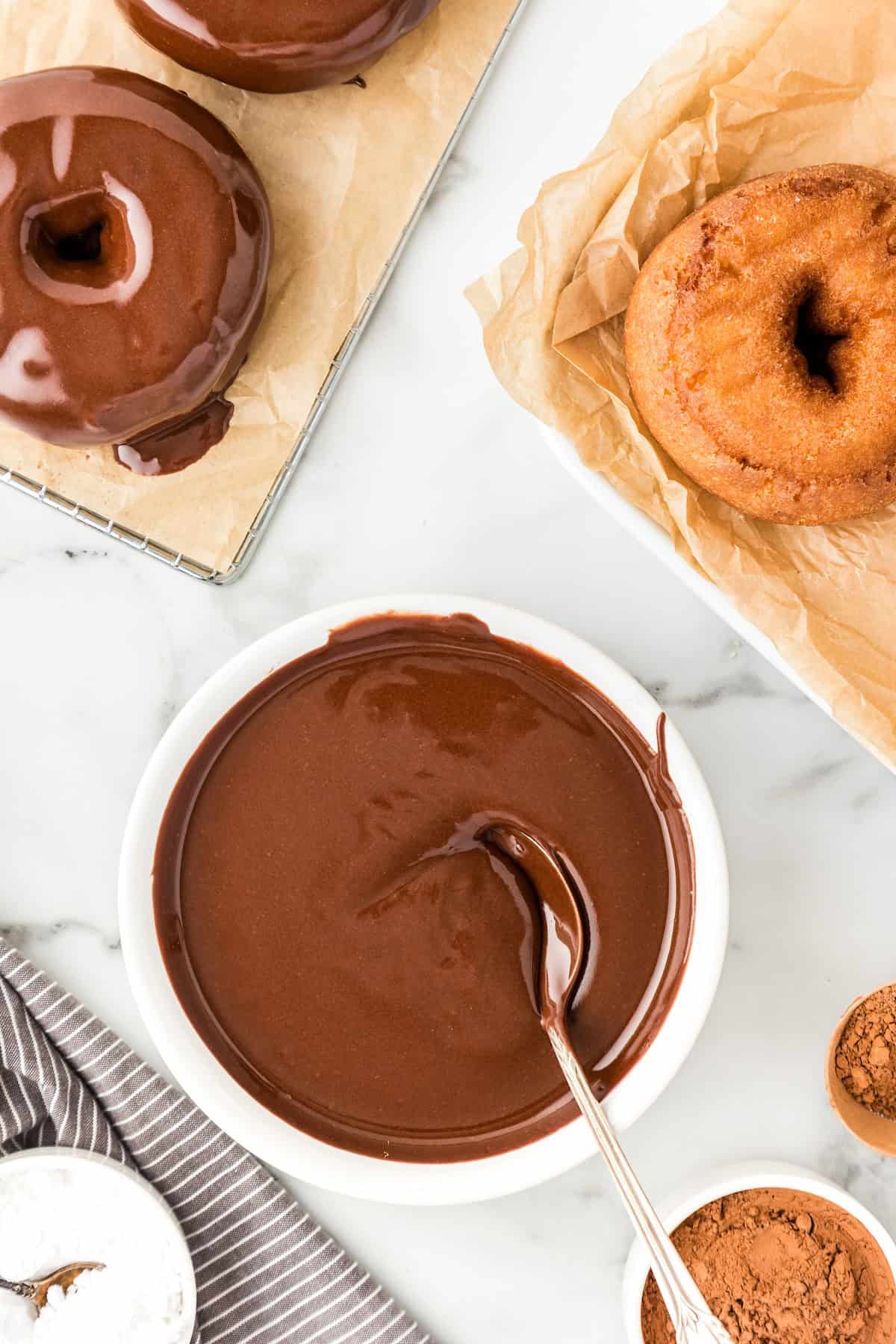 Perfectly rich and smooth, this easy Chocolate Glaze recipe is made with only 4 ingredients: powdered sugar, cocoa powder, milk and vanilla. A bowl of thick chocolate glaze is placed next to donuts.