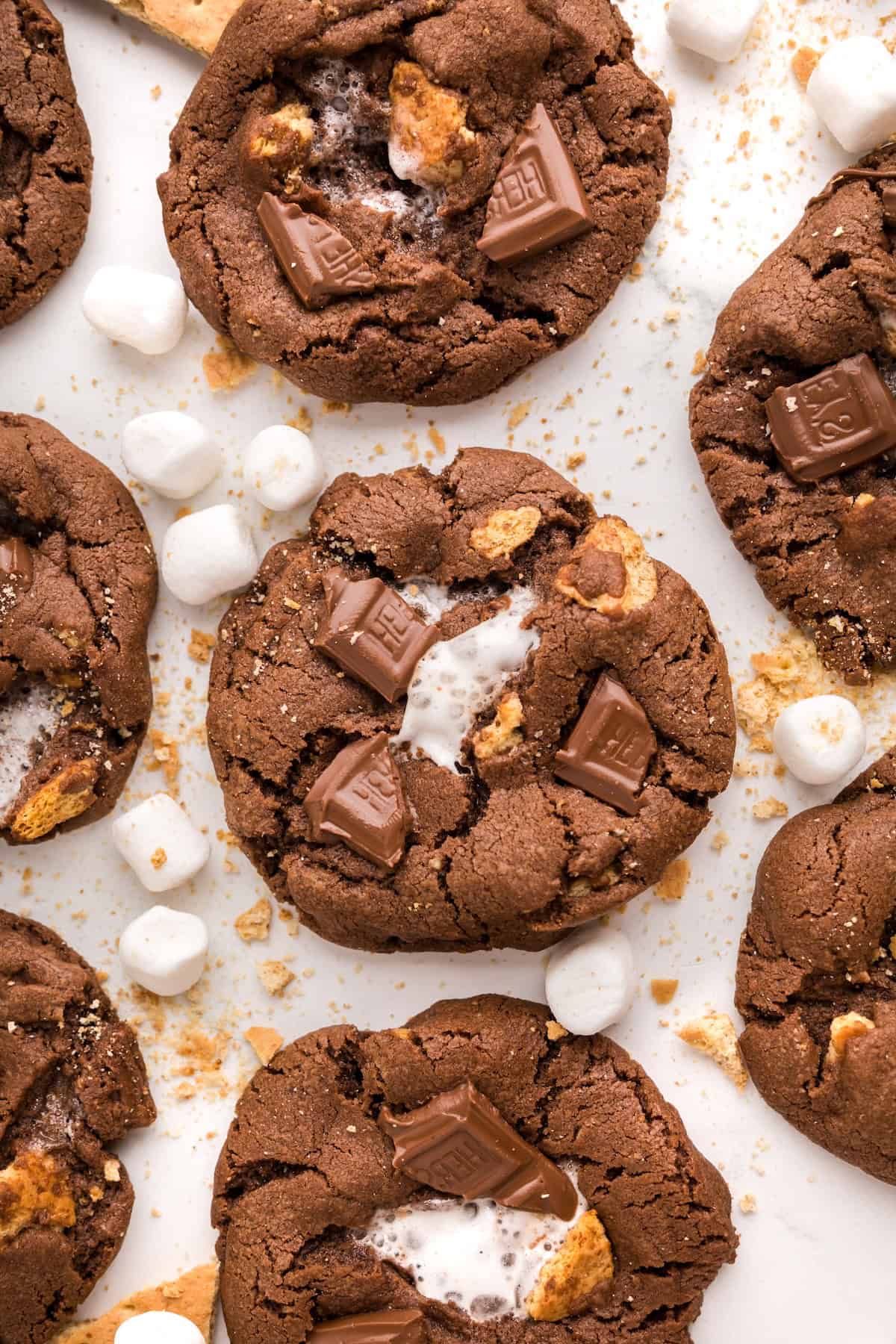 Soft and chewy Chocolate S’mores Cookies are loaded with melty chocolate, graham cracker pieces, and a gooey marshmallow baked in the center! Close-up of chocolate s'mores cookies topped with chunks of chocolate, gooey marshmallow, and bits of graham cracker, surrounded by mini marshmallows and crumbs on a white surface.