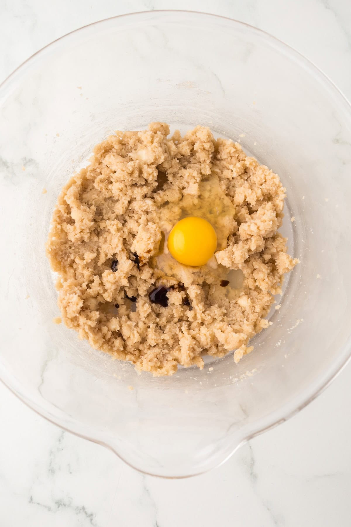 Soft and chewy Chocolate S’mores Cookies are loaded with melty chocolate, graham cracker pieces, and a gooey marshmallow baked in the center! A clear glass mixing bowl with cookie dough mixture and a whole raw egg on top, sitting on a white marble surface.