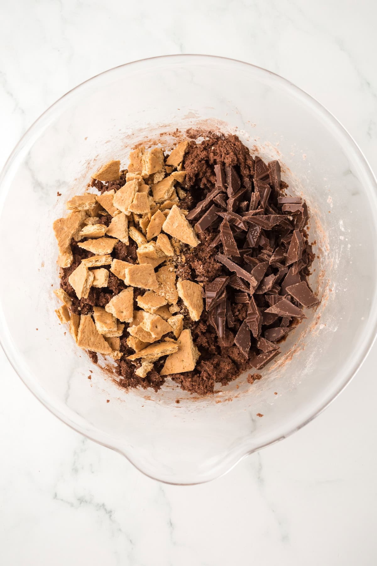 Soft and chewy Chocolate S’mores Cookies are loaded with melty chocolate, graham cracker pieces, and a gooey marshmallow baked in the center! A glass mixing bowl contains chocolate cookie dough topped with chunks of chocolate and pieces of broken graham crackers, sitting on a white marble surface.