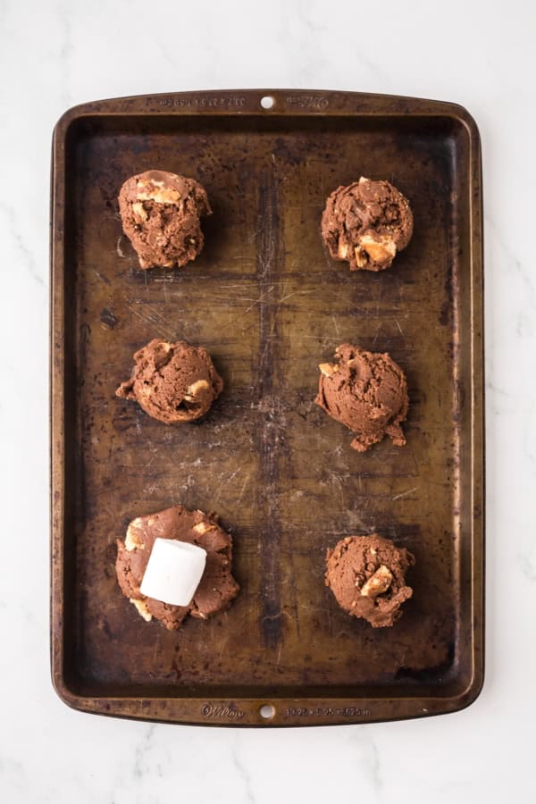 Soft and chewy Chocolate S’mores Cookies are loaded with melty chocolate, graham cracker pieces, and a gooey marshmallow baked in the center! Six scoops of chocolate cookie dough, one topped with a marshmallow, are spaced evenly on a worn metal baking sheet set on a white marble surface.