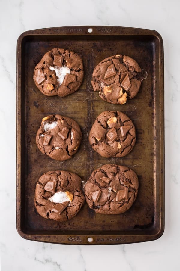 Soft and chewy Chocolate S’mores Cookies are loaded with melty chocolate, graham cracker pieces, and a gooey marshmallow baked in the center! A baking tray with six large chocolate s'mores cookies, some with visible chunks of marshmallow and pieces of chocolate on top, placed on a light marble surface.