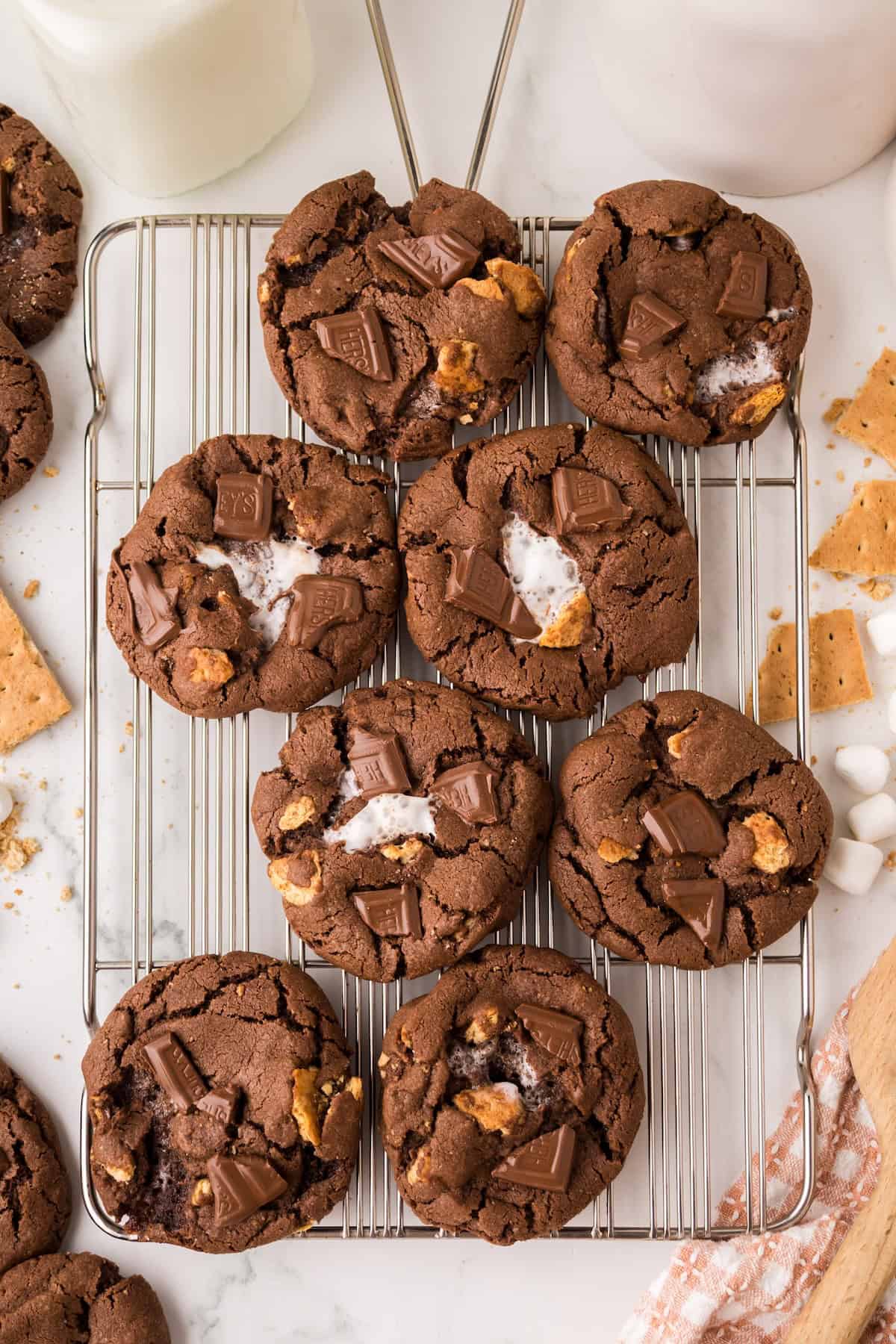 Soft and chewy Chocolate S’mores Cookies are loaded with melty chocolate, graham cracker pieces, and a gooey marshmallow baked in the center! A cooling rack with nine chocolate marshmallow cookies topped with chunks of chocolate, melted marshmallow, and bits of graham cracker. Scattered marshmallows, graham crackers, and a glass of milk are nearby.