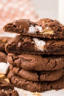 A stack of chocolate s'mores cookies with gooey marshmallow and melted chocolate inside. One cookie on top is broken in half, showing the soft, melty filling. Marshmallow pieces and crumbs are visible nearby.
