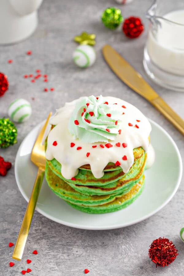 These fluffy Grinch Pancakes with cream cheese frosting are an easy homemade Christmas pancake recipe for a festive Christmas breakfast! Fluffy green grinch pancakes on a plate with cream cheese frosting, green whipped cream and red heart sprinkles on top.