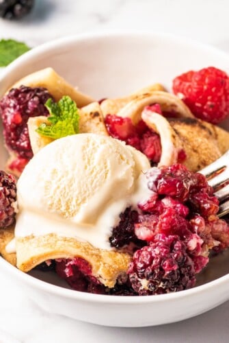 A fork picking up a bite of berry cobbler and vanilla ice cream out of a bowl.