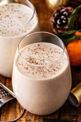 Two glasses of creamy Puerto Rican Coquito sprinkled with ground cinnamon and nutmeg on top, set on a wooden table with a grater, an orange, and a pinecone in the background.