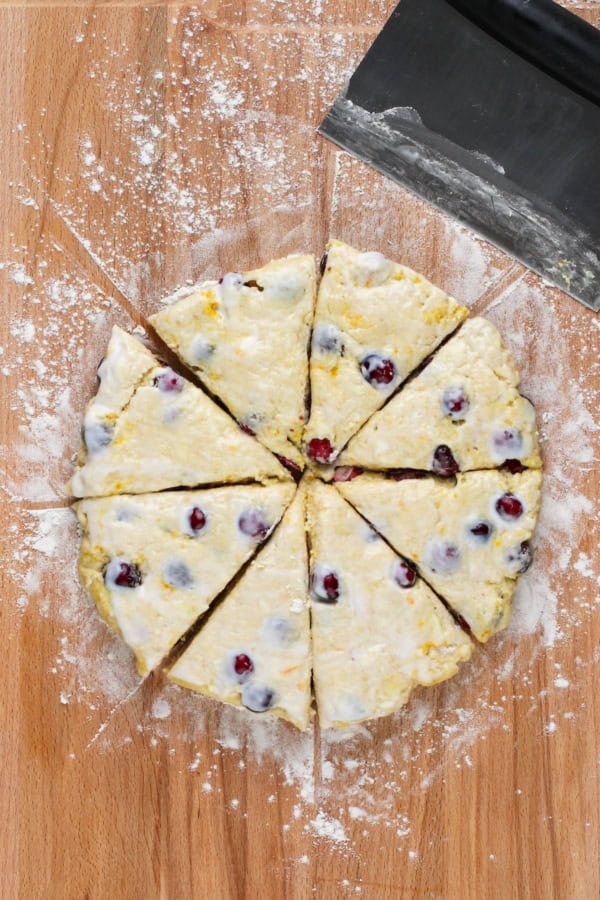 Perfectly dense and flaky, these Cranberry Orange Scones are filled with tart cranberries & fresh orange zest. Easy to make in under an hour! Scone dough has been cut on a wooden surface.