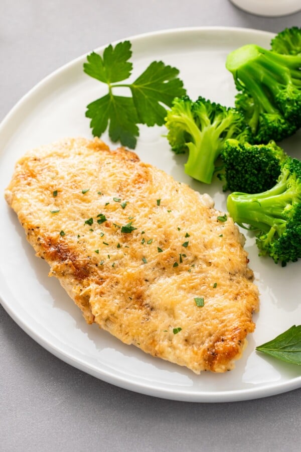 This creamy Garlic Parmesan Chicken recipe is a low-carb dinner that is packed with flavor, but made easy with just 4 simple ingredients! Parsley and steamed broccoli are placed on a plate with a baked parmesan crusted chicken breast.