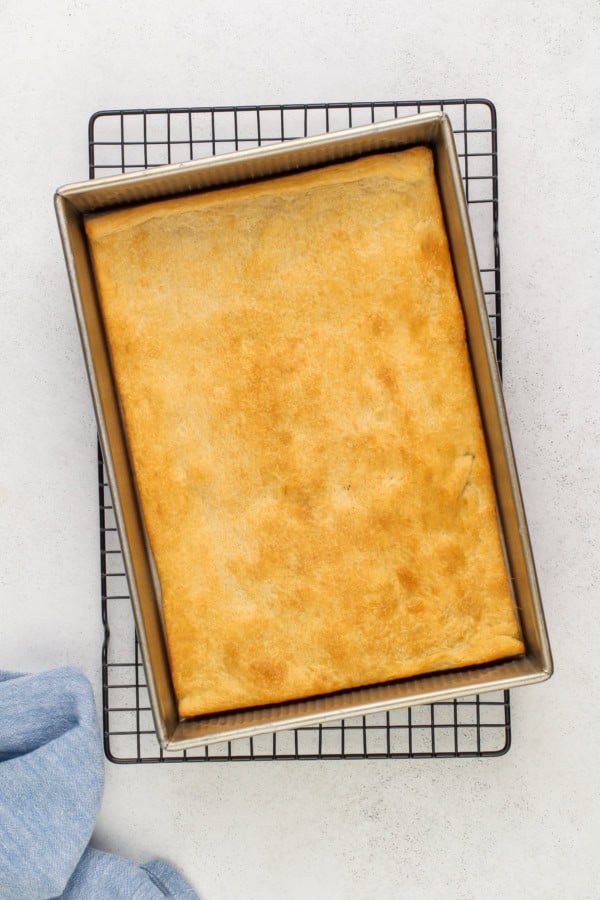 This easy Crescent Roll Veggie Pizza is a classic appetizer made with fresh crunchy veggies and a layer of tangy ranch and cream cheese. A golden-brown rectangular baked crescent dough sits in a metal baking pan on a cooling rack, with a light blue cloth partially visible in the corner.