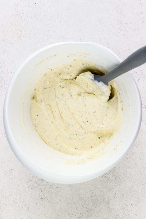 This easy Crescent Roll Veggie Pizza is a classic appetizer made with fresh crunchy veggies and a layer of tangy ranch and cream cheese. A white bowl filled with cream cheese, sour cream, and ranch seasoning mixture with green herb flecks. A gray spoon is partially submerged in the mixture, resting on the side of the bowl.