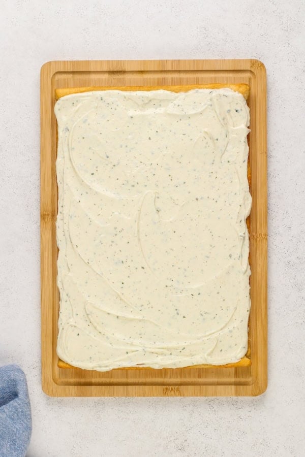 This easy Crescent Roll Veggie Pizza is a classic appetizer made with fresh crunchy veggies and a layer of tangy ranch and cream cheese. A rectangular baked crescent roll dough topped with a layer of creamy ranch and cream cheese mixture, placed on a wooden cutting board against a light background. A blue cloth is partially visible on the lower left corner.