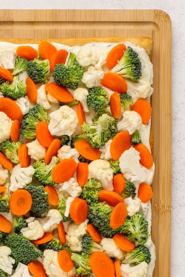 This easy Crescent Roll Veggie Pizza is a classic appetizer made with fresh crunchy veggies and a layer of tangy ranch and cream cheese. A close-up of a rectangular vegetable pizza with crescent roll dough crust topped with a creamy ranch sauce, sliced carrots, broccoli florets, and cauliflower, displayed on a wooden cutting board.
