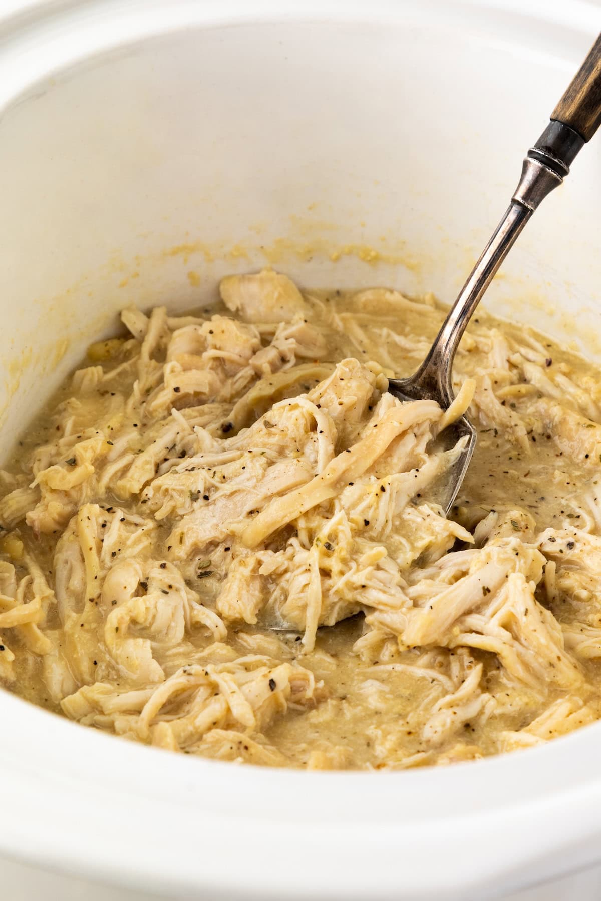 Shredded chicken and gravy inside a white slow cooker, with a metal spoon for serving.