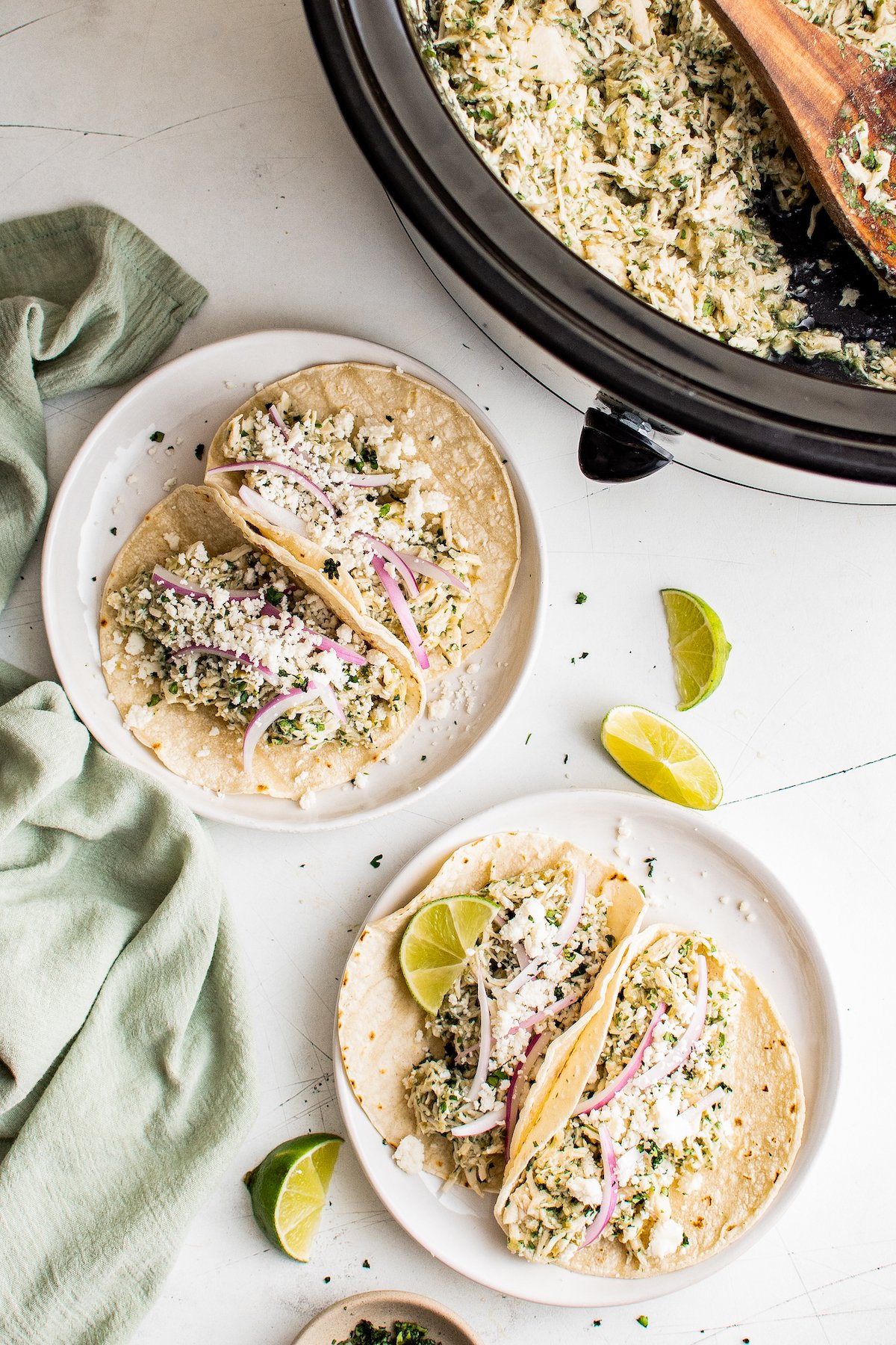 Salsa Verde Chicken Tacos in the Crockpot are an easy Taco Tuesday recipe! Made with tangy, savory salsa verde, these shredded chicken tacos will have everyone in the family fighting for the last one! Plated salsa verde chicken tacos with toppings, next to a slow cooker insert with more salsa verde chicken inside.