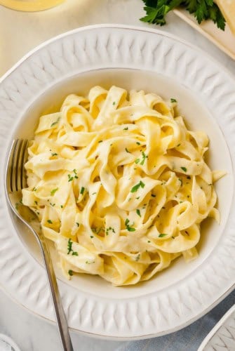 A bowl of creamy fettuccine Alfredo garnished with chopped parsley, served with a fork on a white plate.