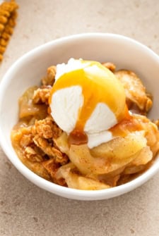 A bowl of apple crisp topped with a scoop of vanilla ice cream and drizzled with caramel sauce, served on a beige surface.