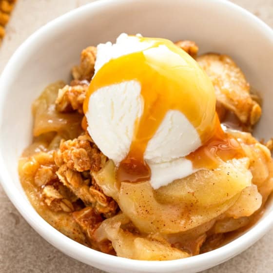A bowl of apple crisp topped with a scoop of vanilla ice cream and drizzled with caramel sauce, served on a beige surface.