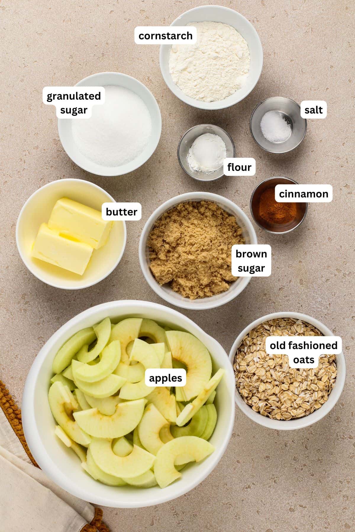 Old‑fashioned Apple Crisp is made with tender, juicy apples baked under a cinnamon‑oat crumble topping. Serve it warm with ice cream! A top-down view of labeled baking ingredients for apple crisp recipe: a bowl of sliced apples, brown sugar, granulated sugar, butter, flour, cornstarch, salt, cinnamon, and old fashioned oats arranged on a light surface.