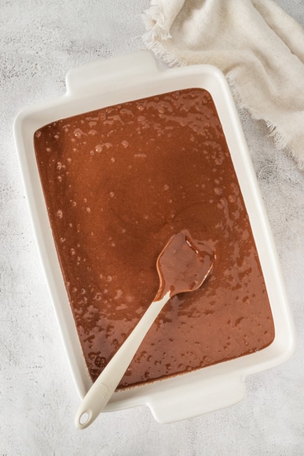 This easy Black Forest Cake recipe is so simple to make with a box of chocolate cake mix, cherry pie filling, and instant chocolate pudding! A white rectangular baking dish filled with chocolate cake batter. A white spatula rests inside, partly covered in the batter. A beige cloth is draped in the top right corner on a light-textured surface.