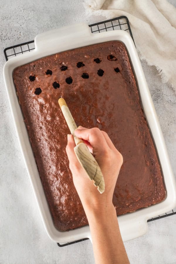 This easy Black Forest Cake recipe is so simple to make with a box of chocolate cake mix, cherry pie filling, and instant chocolate pudding! A white baking dish filled with chocolate cake on a cooling rack. A hand is poking holes in the cake using the handle of a wooden spoon. A beige cloth is partially visible in the background.