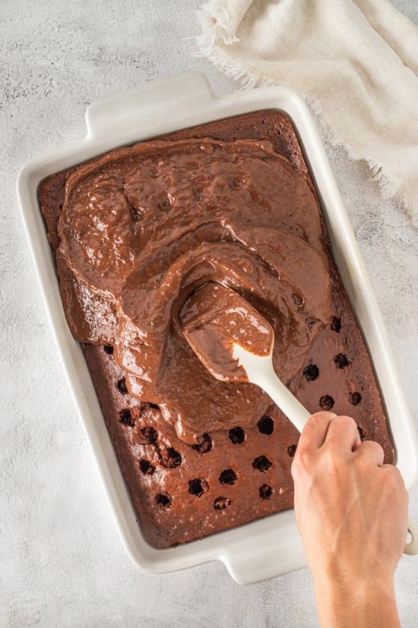 This easy Black Forest Cake recipe is so simple to make with a box of chocolate cake mix, cherry pie filling, and instant chocolate pudding! A persons hand spreading chocolate pudding over a sheet cake with holes using a spatula, in a white rectangular baking dish on a textured light gray surface.