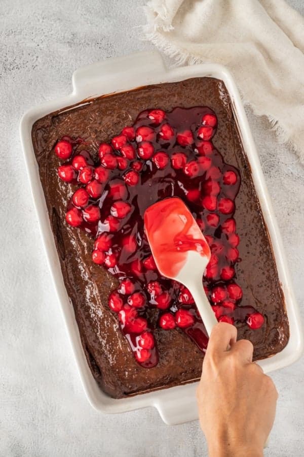 This easy Black Forest Cake recipe is so simple to make with a box of chocolate cake mix, cherry pie filling, and instant chocolate pudding! A hand uses a white spatula to spread cherry pie filling over a freshly baked chocolate cake in a white rectangular dish. The cake is on a light gray surface with a beige cloth nearby.