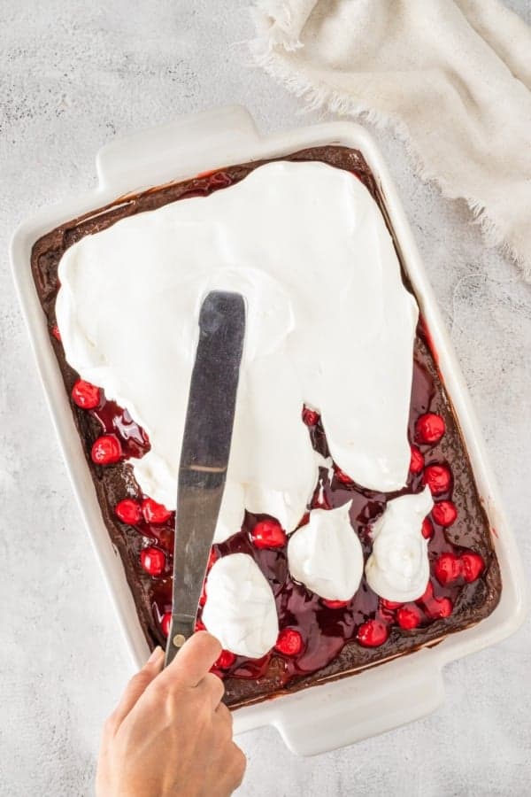 This easy Black Forest Cake recipe is so simple to make with a box of chocolate cake mix, cherry pie filling, and instant chocolate pudding! A hand spreading whipped cream over a cherry-topped chocolate cake using a spatula. The cake is in a rectangular white baking dish.