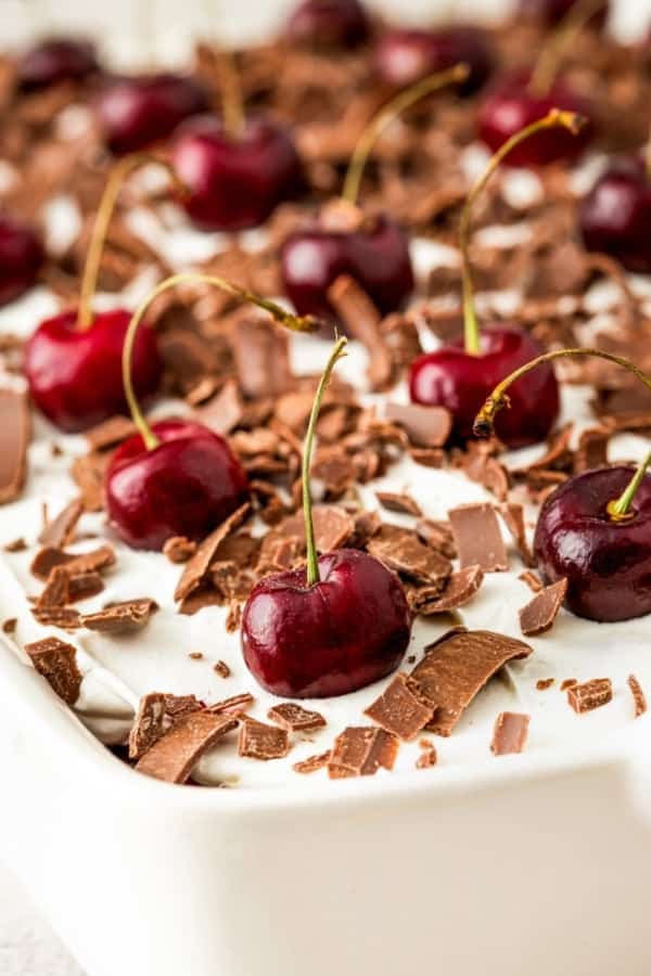 This easy Black Forest Cake recipe is so simple to make with a box of chocolate cake mix, cherry pie filling, and instant chocolate pudding! Close-up of a chocolate cherry cake topped with whipped cream, chocolate shavings, and whole cherries with stems. The cake is in a rectangular white dish, with the cherries and chocolate contrasting against the creamy background.