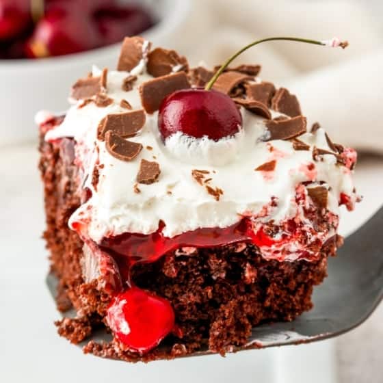 A close-up of a slice of easy Black Forest cake on a spatula. The poke cake has layers of chocolate cake, chocolate pudding, cherry pie filling, and whipped cream. Chocolate shavings and a real cherry adorn the top, with a background of cherries in a bowl.