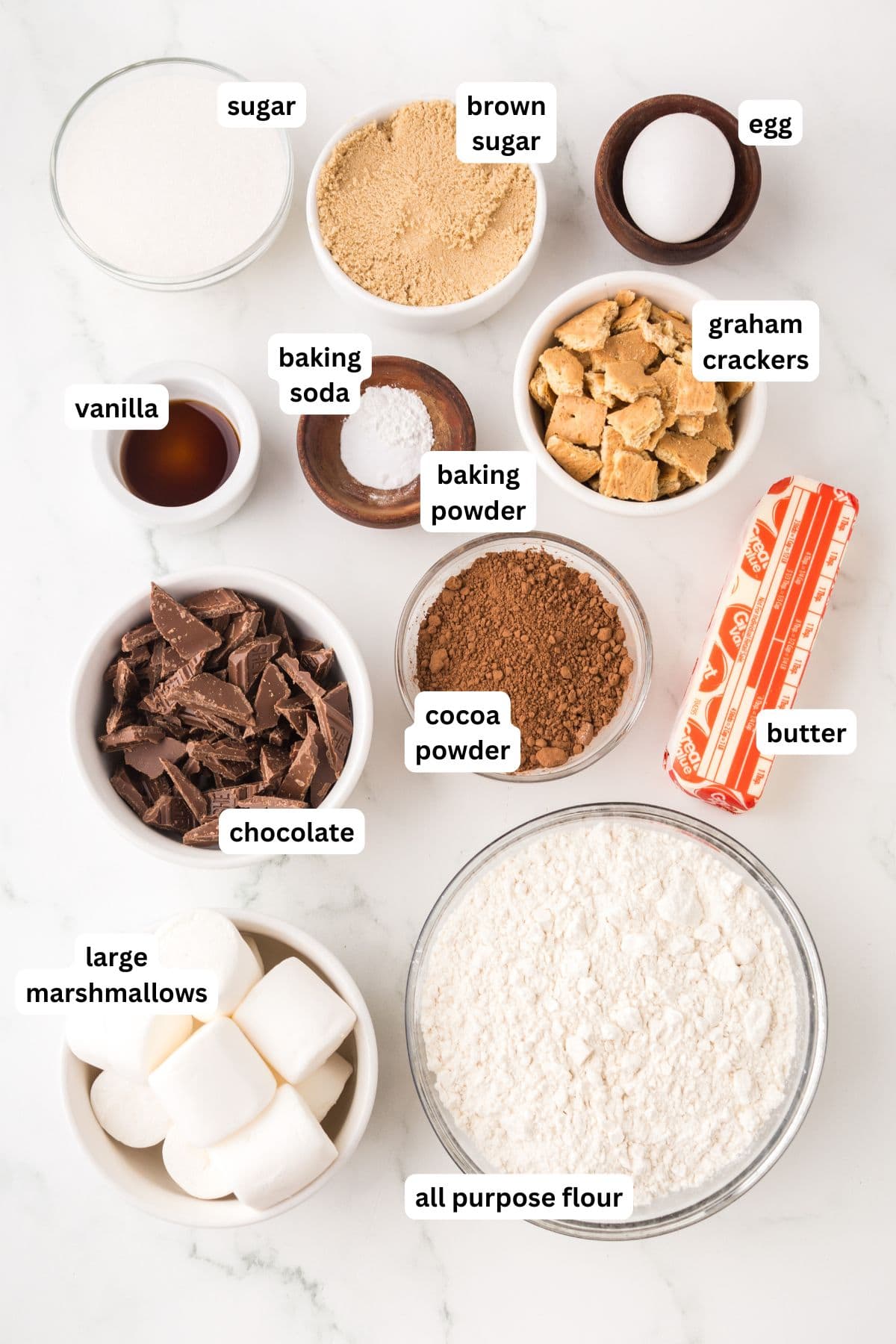 Soft and chewy Chocolate S’mores Cookies are loaded with melty chocolate, graham cracker pieces, and a gooey marshmallow baked in the center! Ingredients for baking chocolate s'mores cookies are arranged on a white surface, each labeled: sugar, brown sugar, egg, baking soda, baking powder, graham crackers, vanilla, chocolate, cocoa powder, butter, all purpose flour, and large marshmallows.