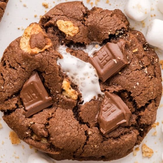 A close-up of a chocolate s'mores cookie with chunks of chocolate, mini marshmallows, and bits of graham cracker, surrounded by more cookies and scattered marshmallows.