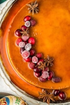 A caramel colored eggnog flan topped with sugared cranberries and star anise, arranged decoratively on one side, served on a glass platter.