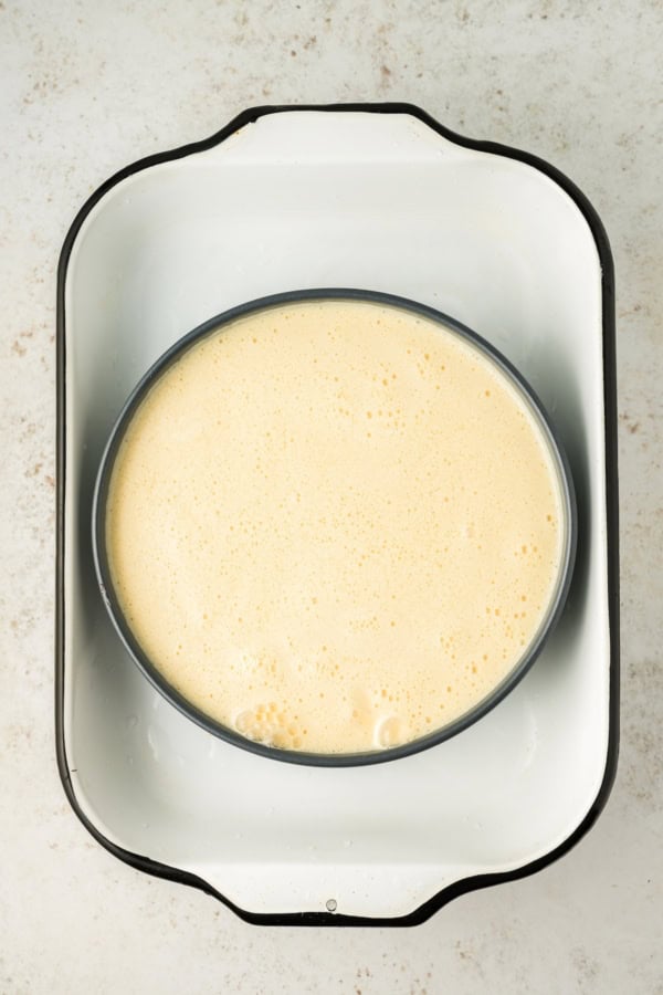 This festive Eggnog Flan is smooth, rich, and creamy, with plenty of nutmeg and rum for that classic spiced holiday flavor. A round baking pan filled with unbaked flan sits inside a larger white roasting pan, ready for a water bath.