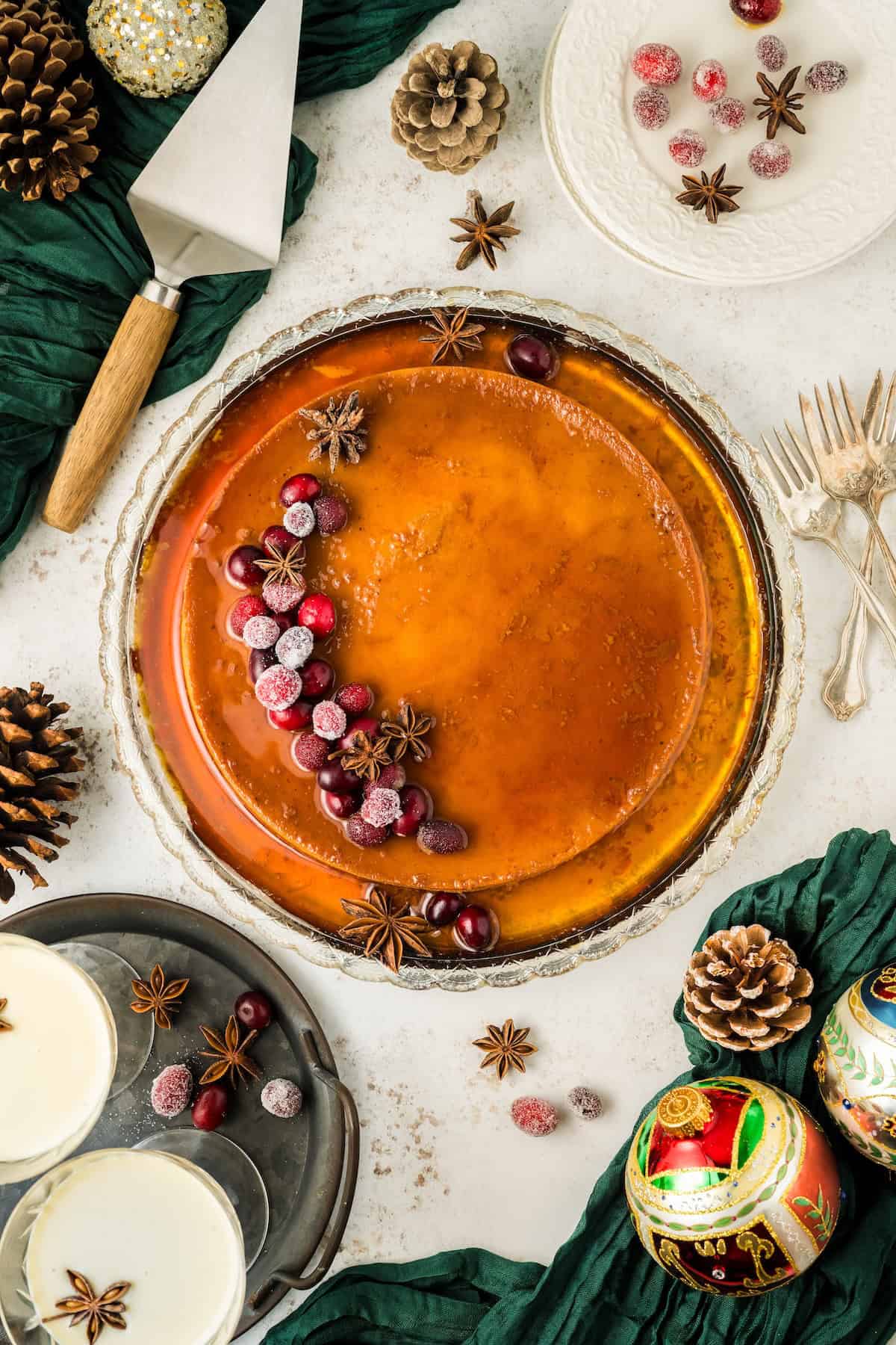 This festive Eggnog Flan is smooth, rich, and creamy, with plenty of nutmeg and rum for that classic spiced holiday flavor. A festive Christmas eggnog flan garnished with cranberries and star anise sits on a table surrounded by glasses of eggnog, festive ornaments, and a pie server.