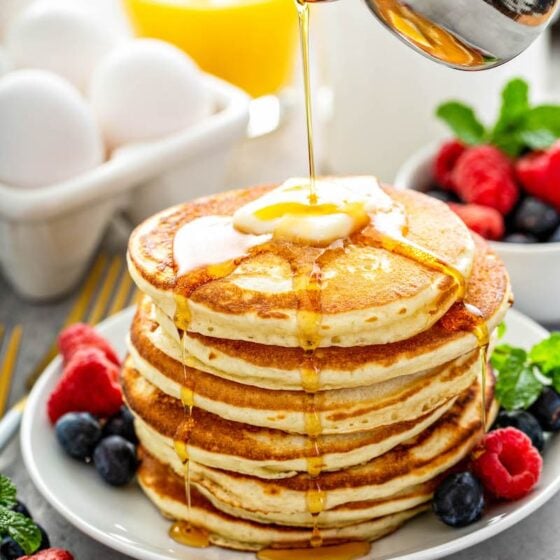 Stack of fluffy pancakes on a white plate with fresh fruit and syrup being poured on top of them.