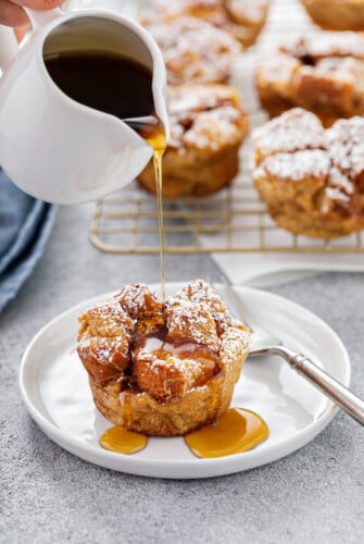 Syrup being poured from a small white pitcher onto a breakfast muffin. More muffins are on a cooling rack in the background.
