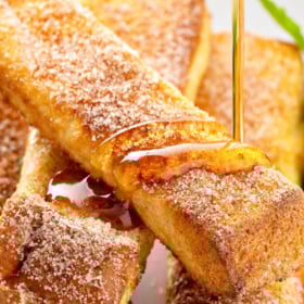 Golden Air Fryer French Toast sticks sprinkled with cinnamon sugar are stacked on a white plate. Syrup is being poured over the top.