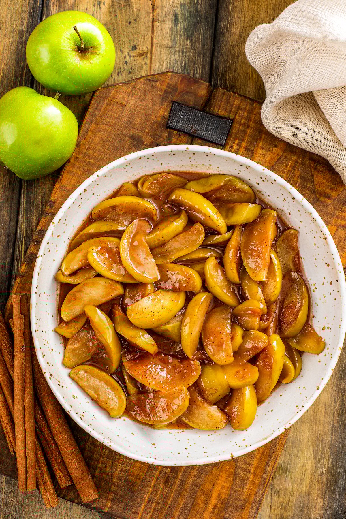 This easy, southern-style Cracker Barrel Fried Apples recipe creates the best tender apples in a comforting cinnamon and brown sugar sauce. Overhead view of a bowl of fried apples with cinnamon sticks and two green apples on the side of the bowl.