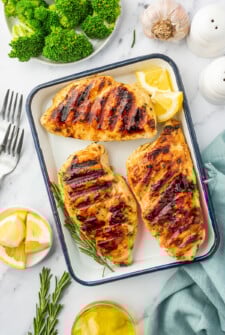 An enamel tray with grilled chicken breast, surrounded by ingredients like broccoli, garlic, lemon wedges, olive oil, and rosemary.