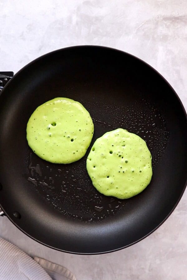 These fluffy Grinch Pancakes with cream cheese frosting are an easy homemade Christmas pancake recipe for a festive Christmas breakfast! Two green grinch pancakes in a skillet being cooked.