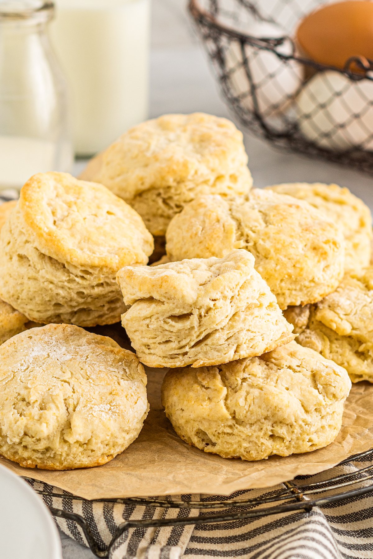 A pile of homemade biscuits on a platter, resting on a blue-and-white striped napkin. A wire basket of eggs and glass bottles of milk are in the background of the shot.