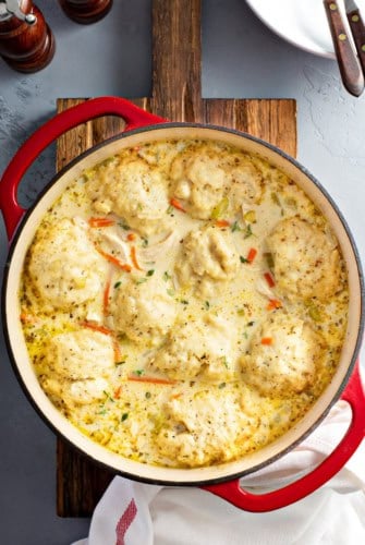 A red Dutch oven filled with creamy homemade chicken and dumplings, featuring fluffy dumplings floating in a thick, herb-speckled broth with visible pieces of carrots and chicken.