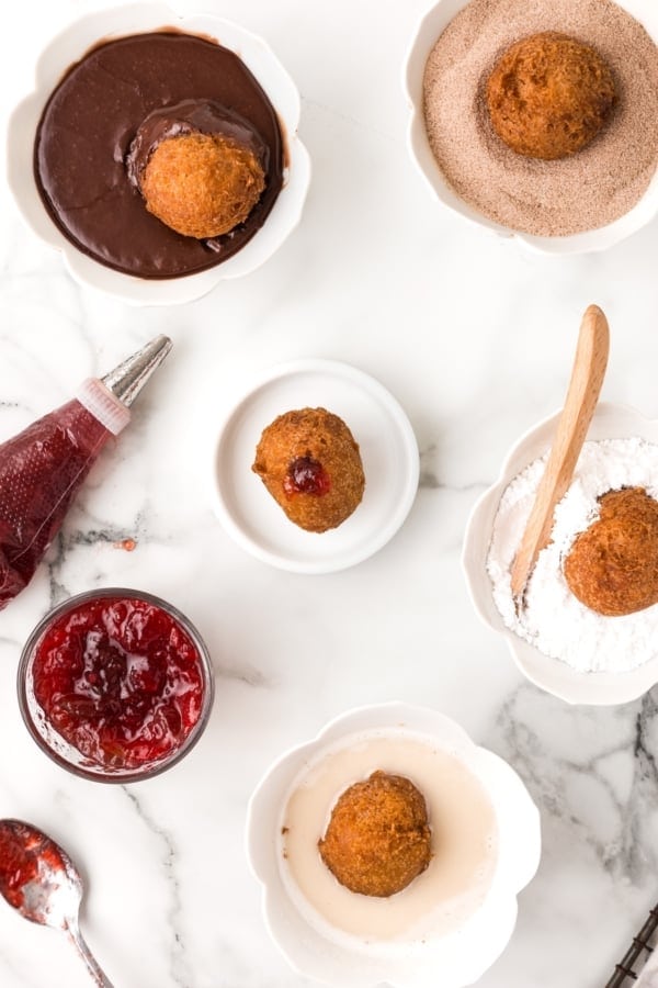 Fresh warm Donut Holes in just 20 minutes — with no yeast or fancy equipment needed! Perfect for dunking, glazing, or stuffing with fillings. Top view of various bowls on a marble surface containing donut holes, each in different coatings: chocolate glaze, cinnamon sugar, and powdered sugar. A jar of red jam, a piping bag, and a spoon with jam are also visible with one donut hole filled with jam.