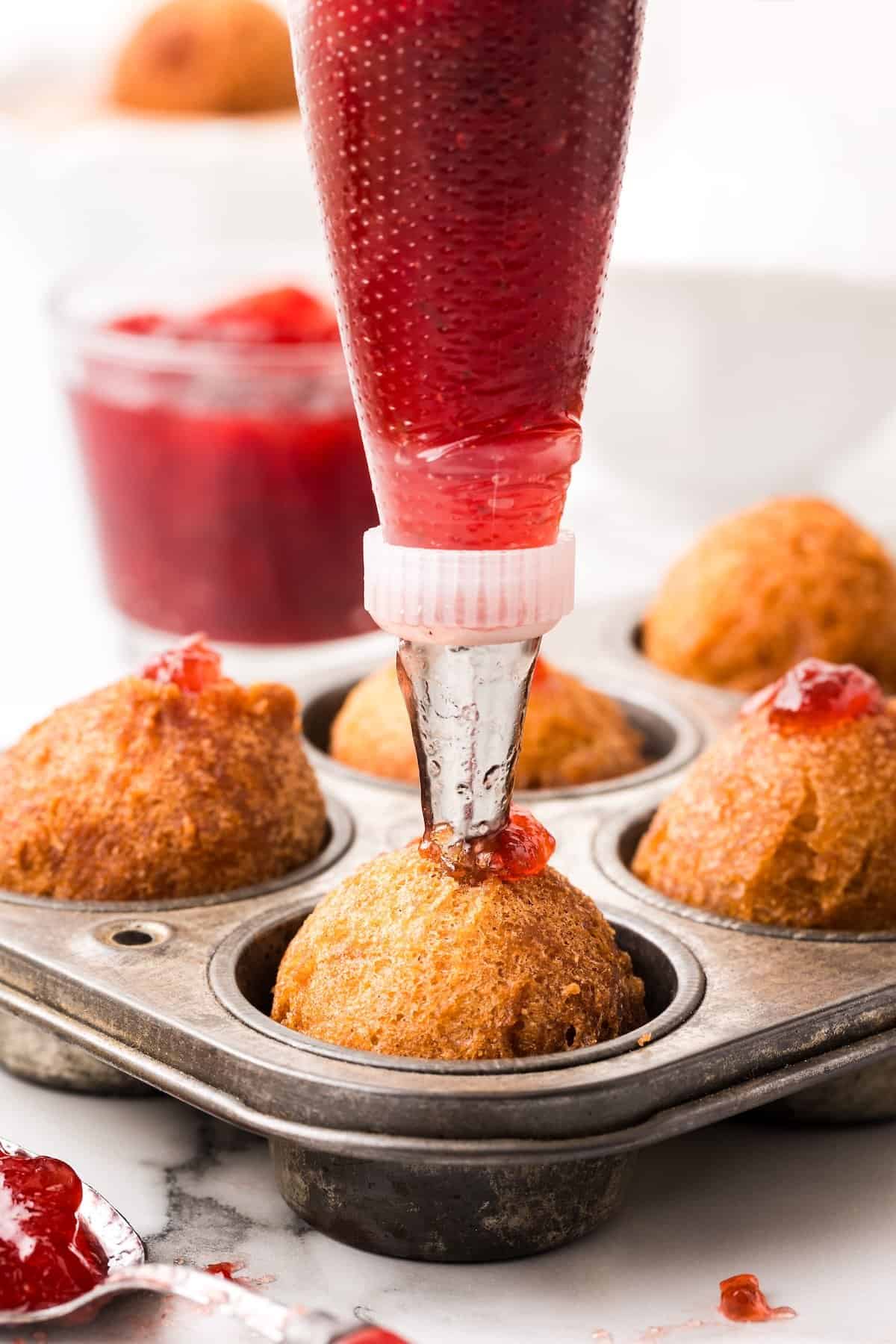 Fresh warm Donut Holes in just 20 minutes — with no yeast or fancy equipment needed! Perfect for dunking, glazing, or stuffing with fillings. A piping bag filled with red jam is injecting the jam into a donut hole in a muffin tin. A jar of red jam and a spoon are in the background on a marble surface.
