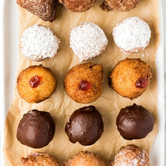An assortment of donut holes on a tray lined with brown paper. The top row is covered in cinnamon sugar, the second row in powdered sugar, the third has a jam filling, the fourth row is coated in chocolate and the bottom row in a traditional donut glaze.