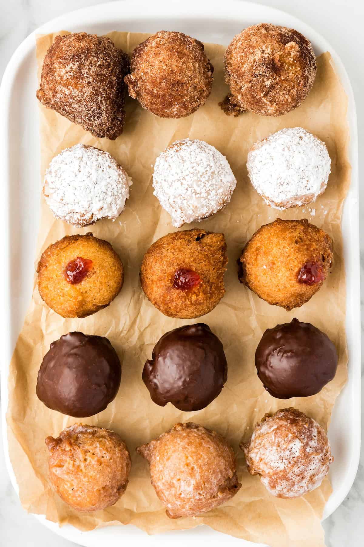 Fresh warm Donut Holes in just 20 minutes — with no yeast or fancy equipment needed! Perfect for dunking, glazing, or stuffing with fillings. An assortment of donut holes on a tray lined with brown paper. The top row is covered in cinnamon sugar, the second row in powdered sugar, the third has a jam filling, the fourth row is coated in chocolate and the bottom row in a traditional donut glaze.